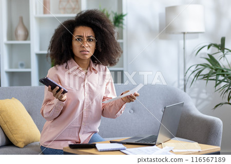Young woman feeling stressed while sitting on couch holding phone and papers. Worried expression suggests financial or work-related issues. Home office setup with laptop and documents on coffee table Young woman feeling stressed while sitting on couch holding phone and papers. Worried expression suggests financial or work-related issues. Home office setup with laptop and documents on coffee table 116567189