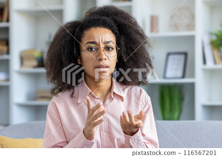 Woman wearing headset sitting at home office, engaging in conversation with frustrated expression on face, gesturing hands. Bookshelves and plants in background, creating professional environment. 116567201
