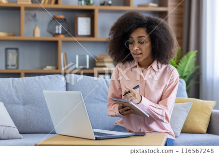 Woman working remotely from home on laptop, taking notes during online meeting. Relaxed professional workspace in living room. Concept of remote work, productivity, technology, and home office. Woman working remotely from home on laptop, taking notes during online meeting. Relaxed professional workspace in living room. Concept of remote work, productivity, technology, and home office. 116567248