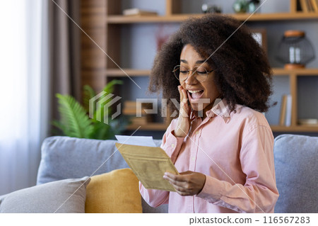 Excited woman in glasses reading letter at home with an expression of surprise and joy. Shows genuine happiness and positive emotions while holding envelope. 116567283