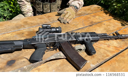 A Kalashnikov assault rifle with a telescopic sight lies on the table in front of the fighter. A Kalashnikov assault rifle with a telescopic sight lies on the table in front of the fighter. 116568531