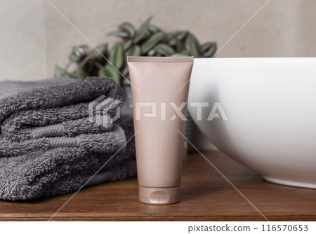 Beige cream squeeze tube near grey towels, plant and basin on wood in bathroom, mockup 116570653