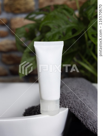 White cream squeeze tube on vessel sink near grey towels and plant close up, cosmetic mockup 116570670