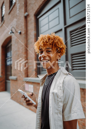Smiling young man holding a smartphone in an urban environment with brick buildings 116571534