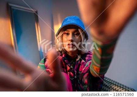Fashionable individual in colorful attire with a blue hat reaching towards the camera 116571560