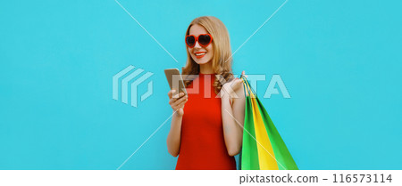 Beautiful happy young woman looking at smartphone with shopping bag in red dress on blue background Beautiful happy young woman looking at smartphone with shopping bag in red dress on blue background 116573114