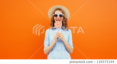 Summer portrait of happy young woman with colorful lollipop, cheerful girl in tourist hat on orange 116573144