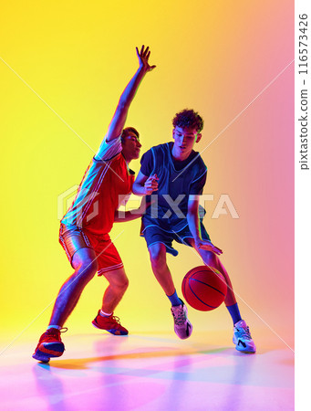 Basketball training with two dedicated athletes in neon light against gradient studio background, emphasizing their agility and determination. Basketball training with two dedicated athletes in neon light against gradient studio background, emphasizing their agility and determination. 116573426