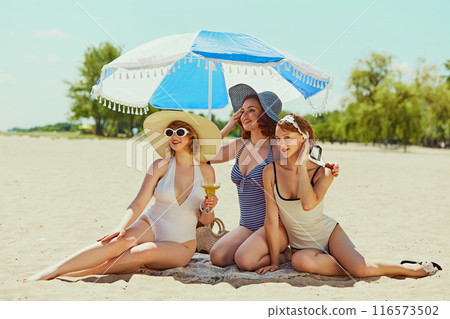 Smiling friends in vintage swimwear, lounging on sand under blue and white umbrella, sharing laughter and drinks. Classic beach day fun. Smiling friends in vintage swimwear, lounging on sand under blue and white umbrella, sharing laughter and drinks. Classic beach day fun. 116573502