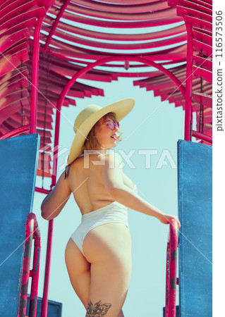 Elegant woman in vintage white swimsuit and wide-brimmed hat, enjoying sunny day on colorful beach lifeguard tower. Classic beach day beauty. 116573506
