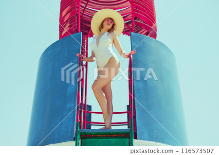 Smiling woman in retro white swimsuit and sun hat, confidently posing on bright red and blue beach lifeguard tower. Timeless summer style. 116573507