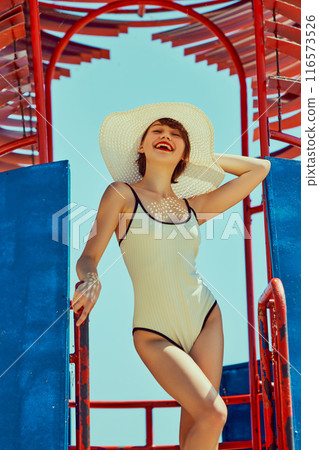 Confident woman in retro white swimsuit with wide sun hat, standing on colorful beach structure, enjoying sunny day. Classic beach fashion. 116573526