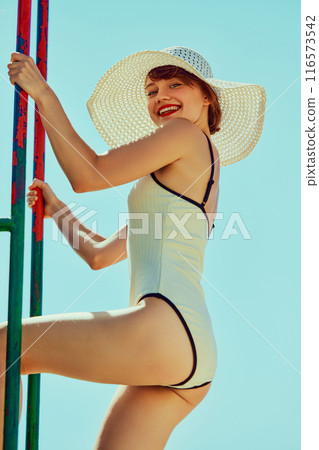 Smiling woman in white one-piece swimsuit with black trim and large sun hat, climbing lifeguard tower against clear blue sky. Vintage summer style. 116573542