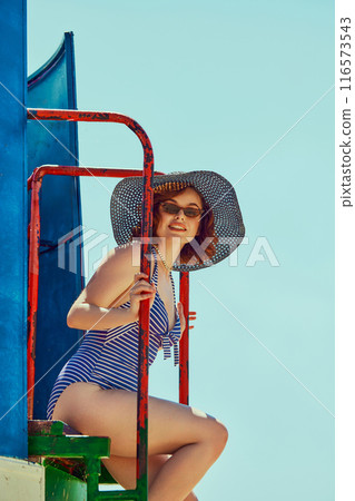Smiling woman wearing blue one-piece swimsuit and wide-brimmed hat, sitting on ladder of lifeguard tower. Retro summer style. Smiling woman wearing blue one-piece swimsuit and wide-brimmed hat, sitting on ladder of lifeguard tower. Retro summer style. 116573543