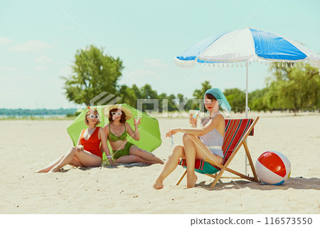 Women in vintage swimsuits and sun hats, enjoying drinks on beach. One sits in chair, and others recline under parasol. Timeless beach day charm. 116573550