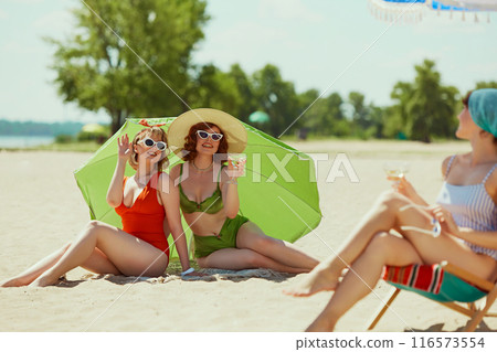 Friends in retro swimwear basking on beach. One lounges under blue umbrella with drink, while other two relax under green parasol. Classic summer fun. 116573554