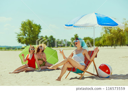 Women in stylish vintage swimsuits, have beach day. One relaxes in beach chair, other two lie under green parasol, all enjoying sunny weather. Women in stylish vintage swimsuits, have beach day. One relaxes in beach chair, other two lie under green parasol, all enjoying sunny weather. 116573559