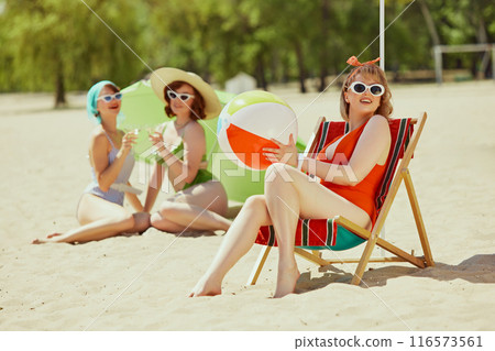 Group of women in vintage swimsuits, sipping drinks and soaking up sun on beach. One sits in beach chair, others lie on sand with parasol. 116573561