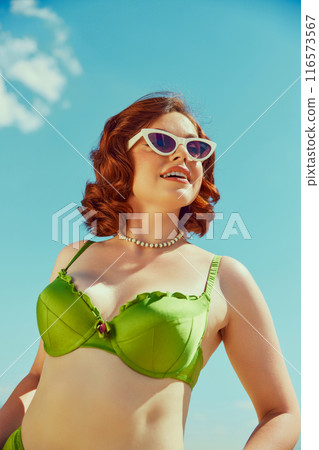 Smiling woman in green retro bikini, pearl necklace, white sunglasses, enjoying sunny day at beach. Classic summer fun. 116573567