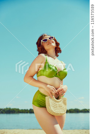 Woman in vintage green bikini, white sunglasses, and pearl necklace, holding straw bag, posing happily under bright blue sky. Classic beach style. Woman in vintage green bikini, white sunglasses, and pearl necklace, holding straw bag, posing happily under bright blue sky. Classic beach style. 116573589