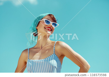 Woman in blue and white striped swimsuit, smiling broadly, under bright blue sky, embodying retro summer joy. 116573598