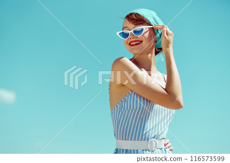 Cheerful woman in striped swimsuit and blue headscarf, wearing white cat-eye sunglasses, basking in sunshine. Retro beach style. Cheerful woman in striped swimsuit and blue headscarf, wearing white cat-eye sunglasses, basking in sunshine. Retro beach style. 116573599