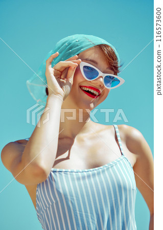 Happy woman in blue striped swimsuit, white belt, and vintage sunglasses, enjoying sunny day. Retro summer fashion. Happy woman in blue striped swimsuit, white belt, and vintage sunglasses, enjoying sunny day. Retro summer fashion. 116573600