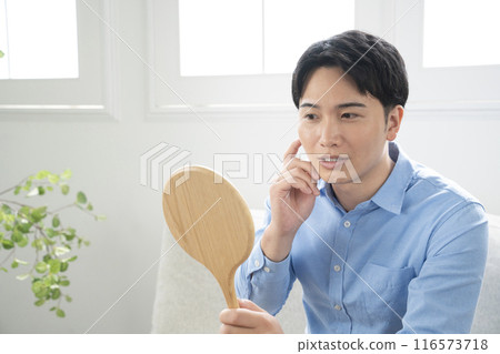 A man in his twenties sitting on a sofa in the living room and looking at a hand mirror 116573718