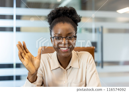Smiling woman waving during video call in modern office. Friendly professional engaging in remote communication. Positive connection, casual yet professional ambiance, indicating ease and 116574092