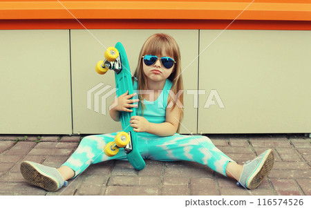Stylish little girl child sitting on the ground with skateboard, happy kid posing on city street 116574526