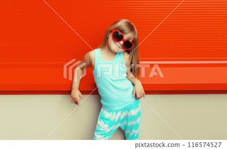 Happy kid girl in red heart shaped glasses, stylish little child posing on city street on background 116574527