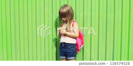 Little girl child standing with backpack on green background on city street Little girl child standing with backpack on green background on city street 116574528
