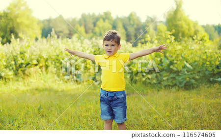 Happy little boy child enjoying warm weather standing on the grass in summer green park 116574605
