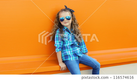 Happy kid girl with curly hair, stylish little child posing in the city on orange wall 116574616