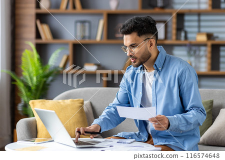 Young man working from home with laptop and documents in modern living room. Casual setting with wooden shelves, plants, and cozy sofa. Focused on work and productivity. 116574648