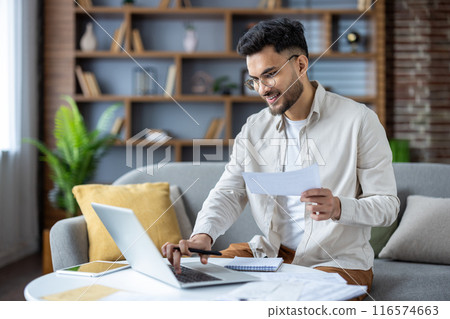 Indian young man working with documents and bills at home, sitting on sofa. holds papers and uses a laptop. Indian young man working with documents and bills at home, sitting on sofa. holds papers and uses a laptop. 116574663