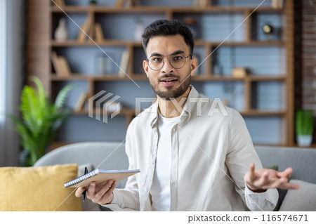 A close-up portrait of a young Muslim man in glasses and a shirt who is at home, holding a pen and a notebook and waving his arms inexplicably to the camera. 116574671