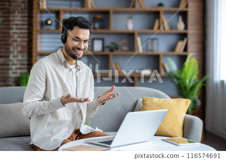Smiling young Indian man sitting on sofa at home wearing headset, talking on video call, gesturing with hands. 116574691