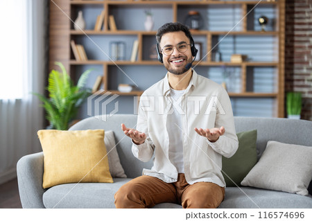 Smiling man wearing headset engaged in video call, sitting comfortably on sofa in home office. Person enjoying virtual meeting from cozy living room environment, expressing positive emotions. 116574696