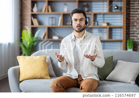 Worried young Indian man is at home sitting on the sofa in a headset talking nonsense, explaining, discussing to the camera, waving his arms. Worried young Indian man is at home sitting on the sofa in a headset talking nonsense, explaining, discussing to the camera, waving his arms. 116574697