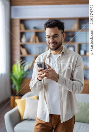 Vertical shot of a young Indian man at home, standing in a room and smiling while using a mobile phone. Vertical shot of a young Indian man at home, standing in a room and smiling while using a mobile phone. 116574743