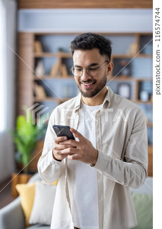 A cheerful Indian man is engaged with his smartphone, enjoying a relaxed time in his modern living room with stylish interior design. He represents a cozy domestic scene in a multi-ethnic context. A cheerful Indian man is engaged with his smartphone, enjoying a relaxed time in his modern living room with stylish interior design. He represents a cozy domestic scene in a multi-ethnic context. 116574744