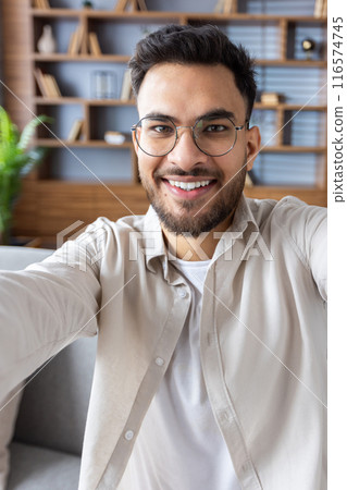 Smiling man at home office making video call using phone, wearing glasses and casual clothes. Bookshelf and plants in background create cozy work environment. Smiling man at home office making video call using phone, wearing glasses and casual clothes. Bookshelf and plants in background create cozy work environment. 116574745