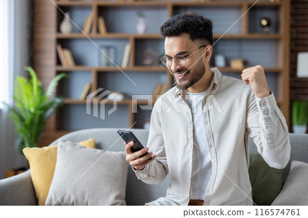 Man sitting on couch expressing excitement while looking at smartphone. Modern home interior with wooden shelves and plants in background. Concept of success, happiness, and achievement. 116574761