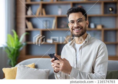 Smiling young man using smartphone in modern living room, seated on comfortable sofa. Bright and cozy home office setting, featuring wooden bookshelf and green plant background. Smiling young man using smartphone in modern living room, seated on comfortable sofa. Bright and cozy home office setting, featuring wooden bookshelf and green plant background. 116574763