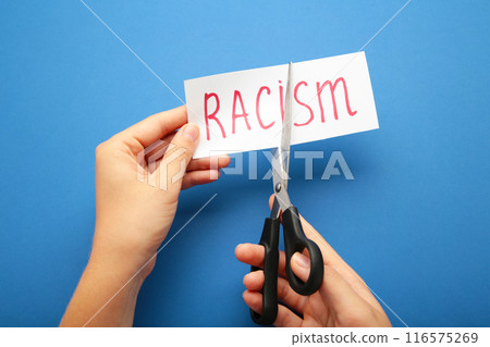 Hand holding card with the text racism. Top view of black scissors and cutted paper card with word Racism on the blue background. Hand holding card with the text racism. Top view of black scissors and cutted paper card with word Racism on the blue background. 116575269