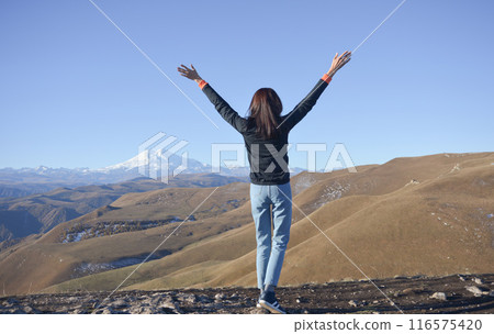 A young brunette woman with her hands raised up against the backdrop of the snow-capped Mount Elbrus A young brunette woman with her hands raised up against the backdrop of the snow-capped Mount Elbrus 116575420