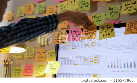 Male professional adds colorful notes to his computer screen amidst a vibrant array of papers, concept of managing time and planning 116575963
