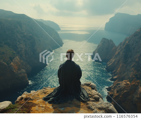 Man dark cloak sits meditating edge cliff overlooking fjord cliffs soft light dawn back view 116576354