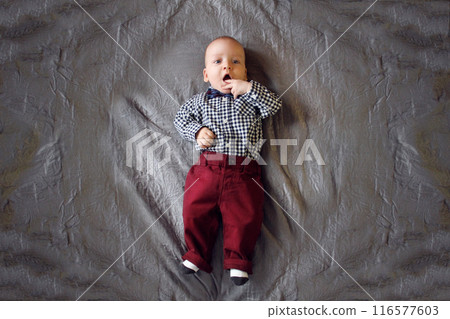 A baby in a plaid shirt and maroon pants lies on the bed A baby in a plaid shirt and maroon pants lies on the bed 116577603